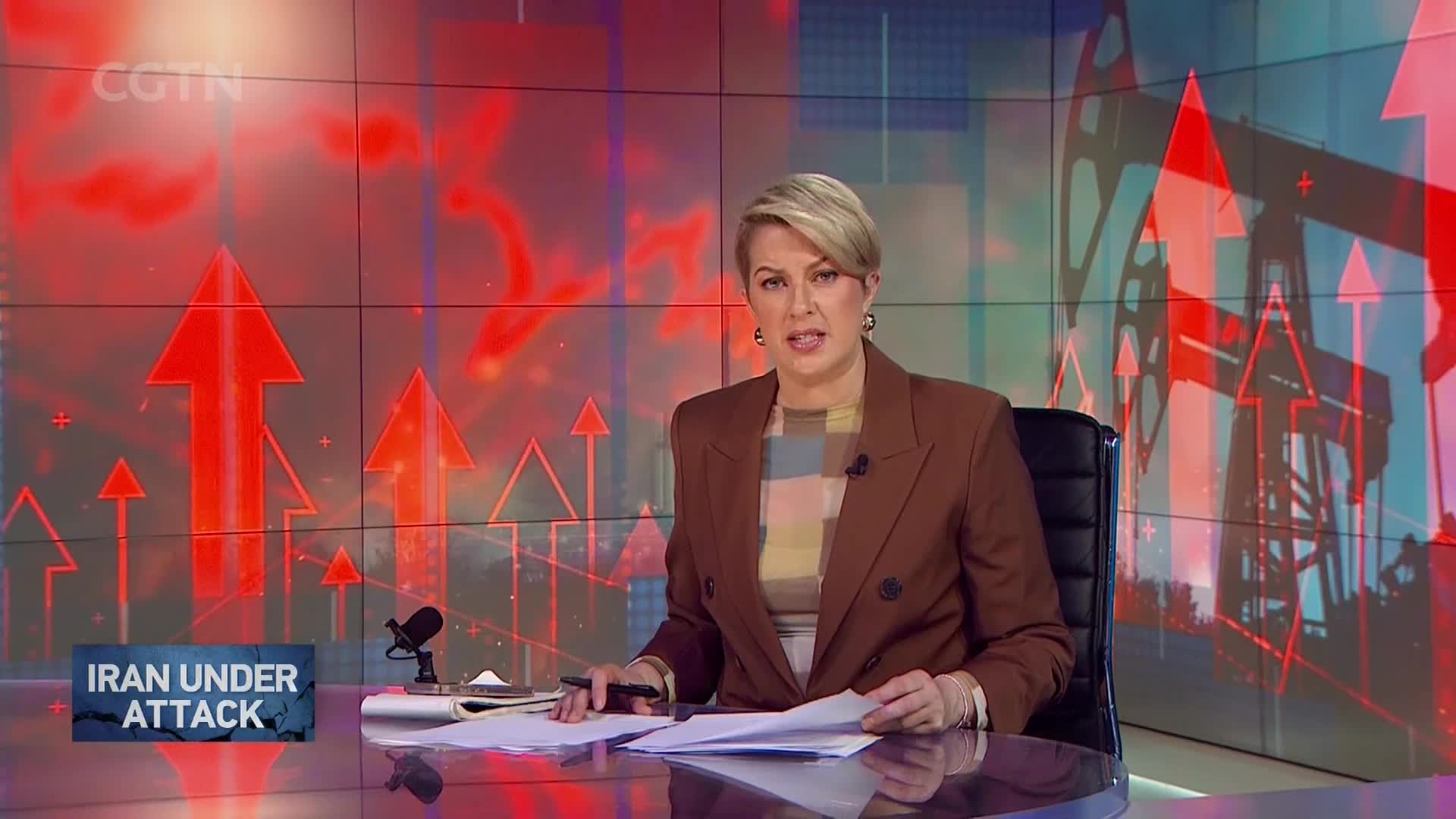 A woman in a brown blazer sits at a desk, reading from papers. Behind her, the CGTN screen displays red arrows pointing upward and images of oil derricks.