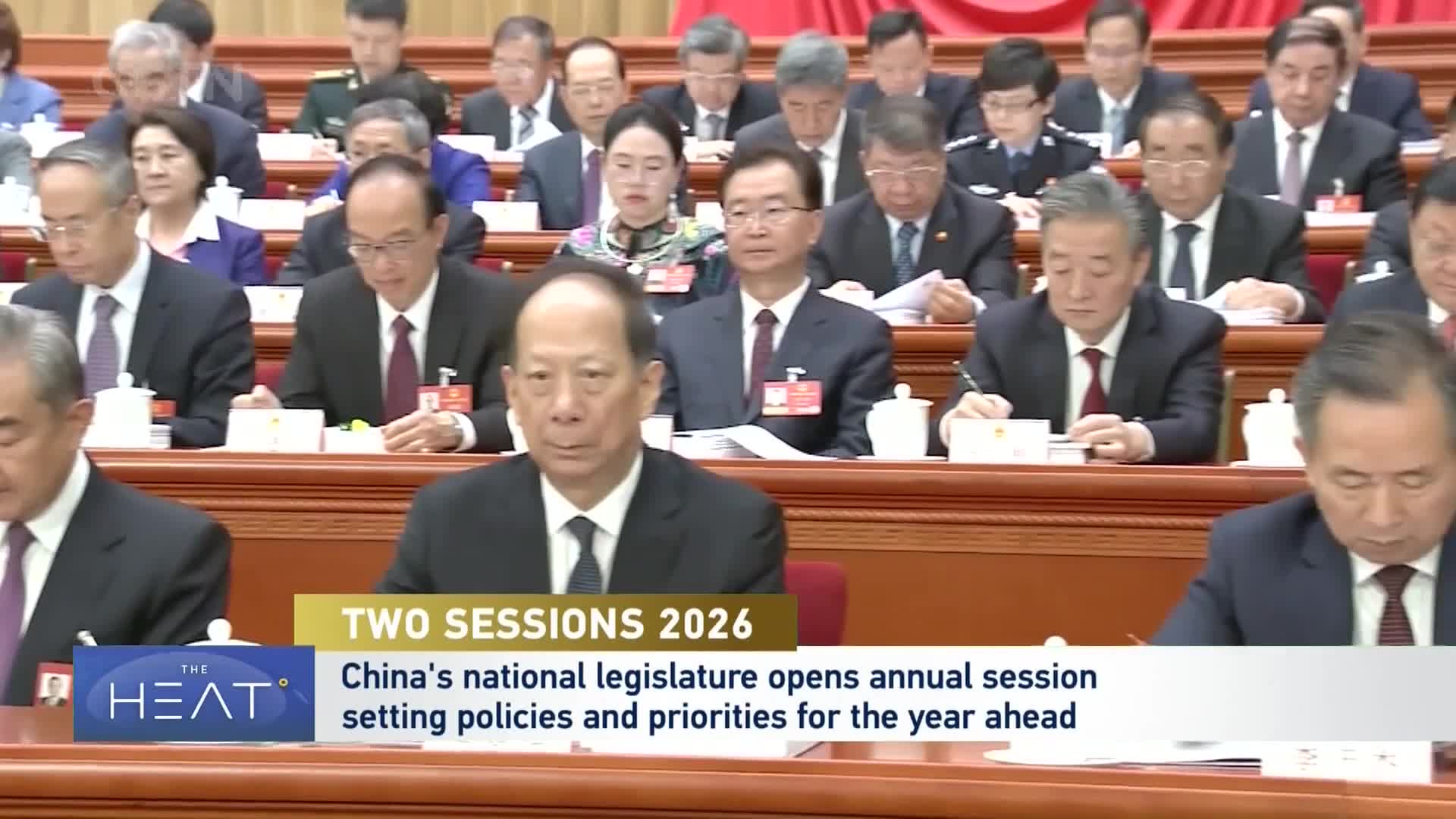 Men in dark suits and ties sit at long tables, some looking down at papers. A banner at the bottom announces "Two Sessions 2026," detailing China's national legislature opening its annual session.