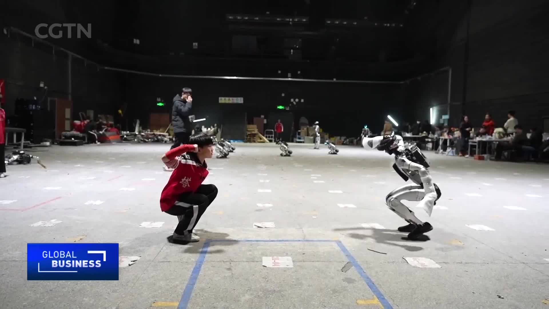 A young man in a red jacket and black pants crouches low, facing a robot in a white suit. The robot mirrors his stance, its metallic limbs bent at the knees.