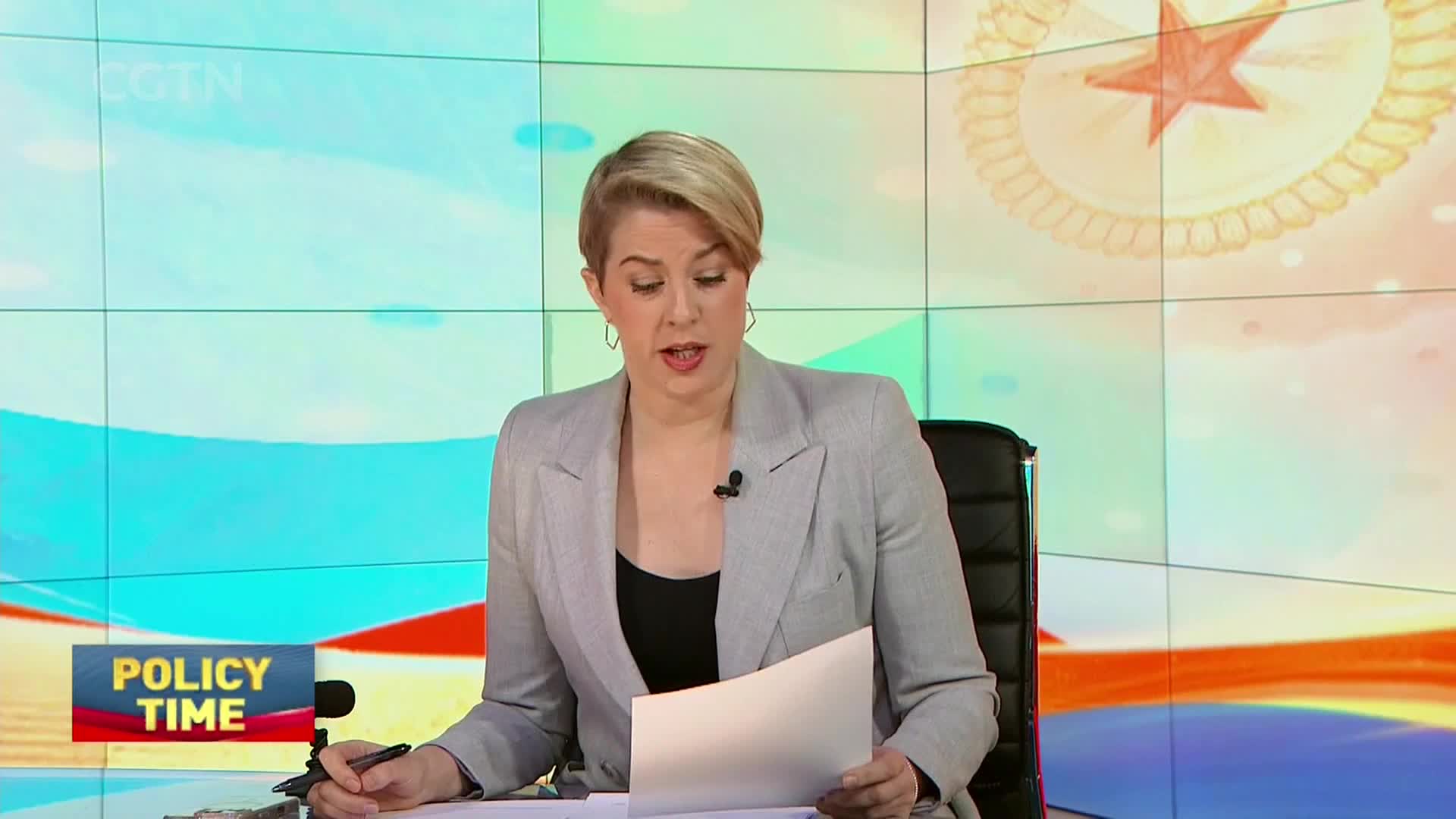 A woman in a gray blazer speaks into a microphone, holding a paper. A graphic for "POLICY TIME" appears in the lower left corner.
