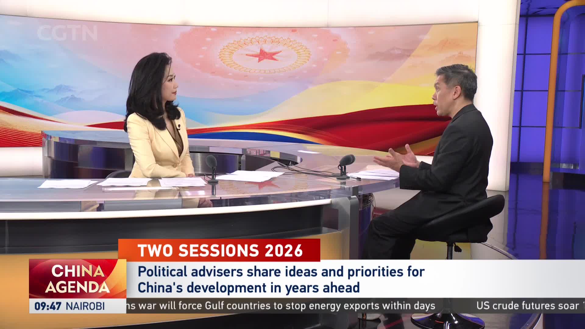 A woman in a light yellow jacket and a man in a dark suit sit across a desk, engaged in conversation. Behind them, a screen displays "CGTN" and graphics related to China's agenda.