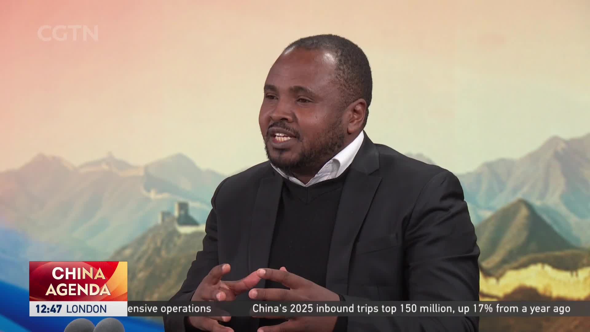A man in a dark suit speaks animatedly, his hands gesturing as he talks. Behind him, a digital backdrop shows the Great Wall snaking over rugged mountains, a scene that CGTN often features.