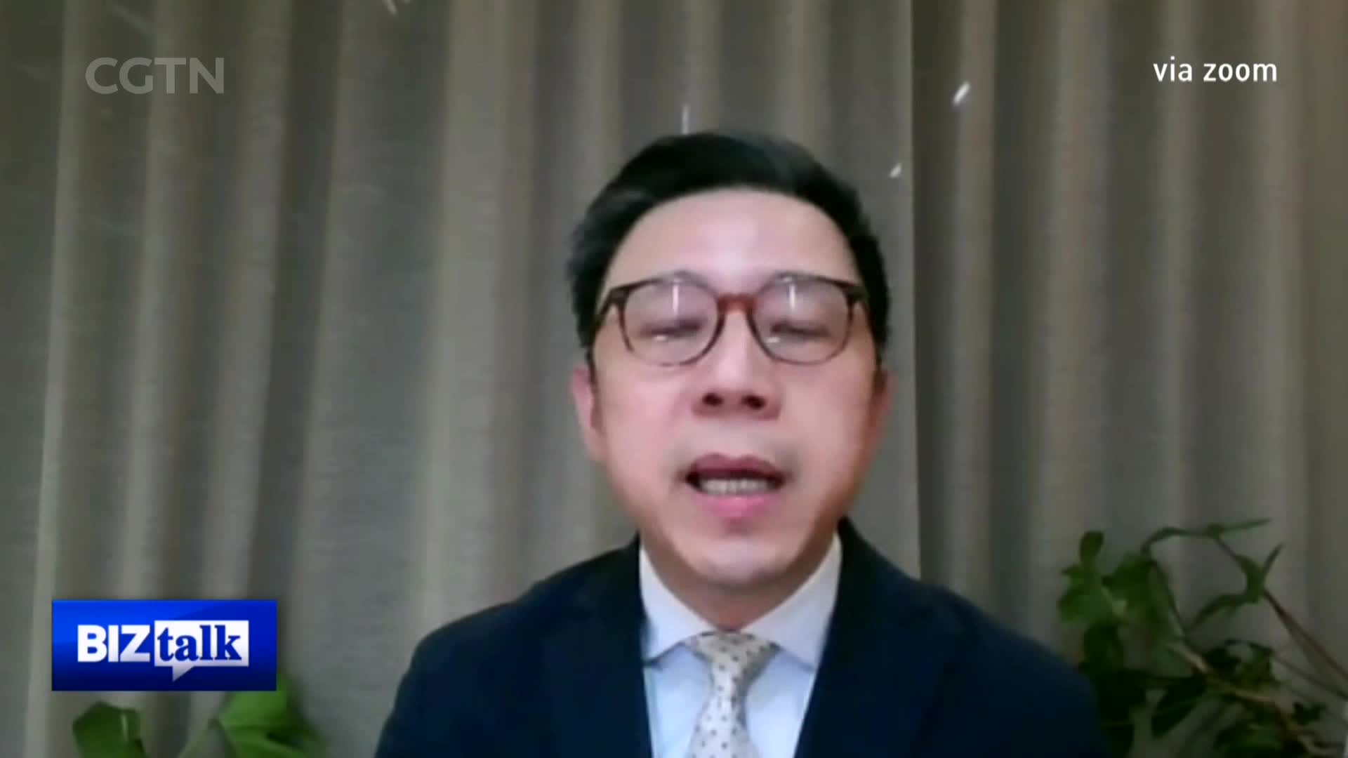 A man in a suit and tie speaks directly to the camera, his glasses reflecting the light. He's participating in a CGTN "BizTalk" segment, appearing via Zoom from China.