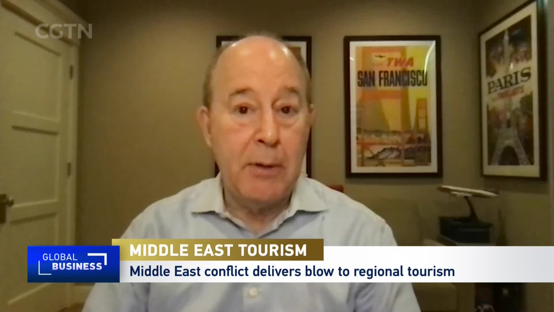 A man speaks on a video call, his image framed by a "Global Business" graphic and a CGTN logo. Behind him, vintage travel posters for San Francisco and Paris hang on the wall.