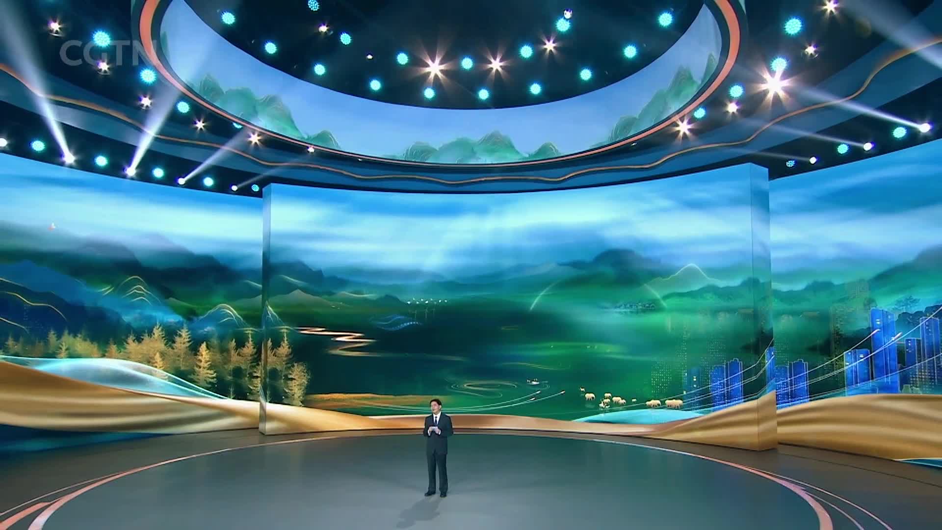 A man in a dark suit stands center stage, facing a massive curved screen displaying a serene Chinese landscape. Lights shine down from above, illuminating the polished floor of the studio.