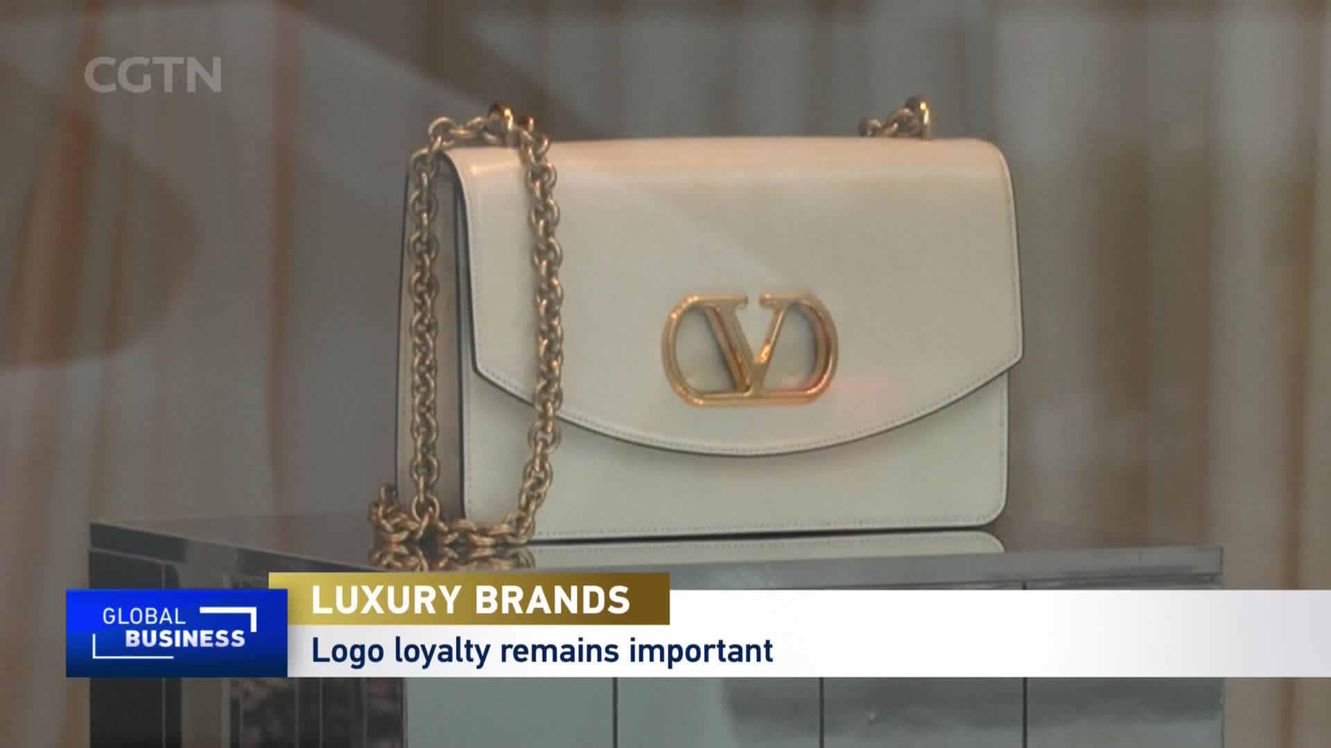 A white leather handbag with a gold chain strap sits on a reflective surface. The CGTN "Global Business" chyron indicates this is a report on luxury brands and logo loyalty in China.