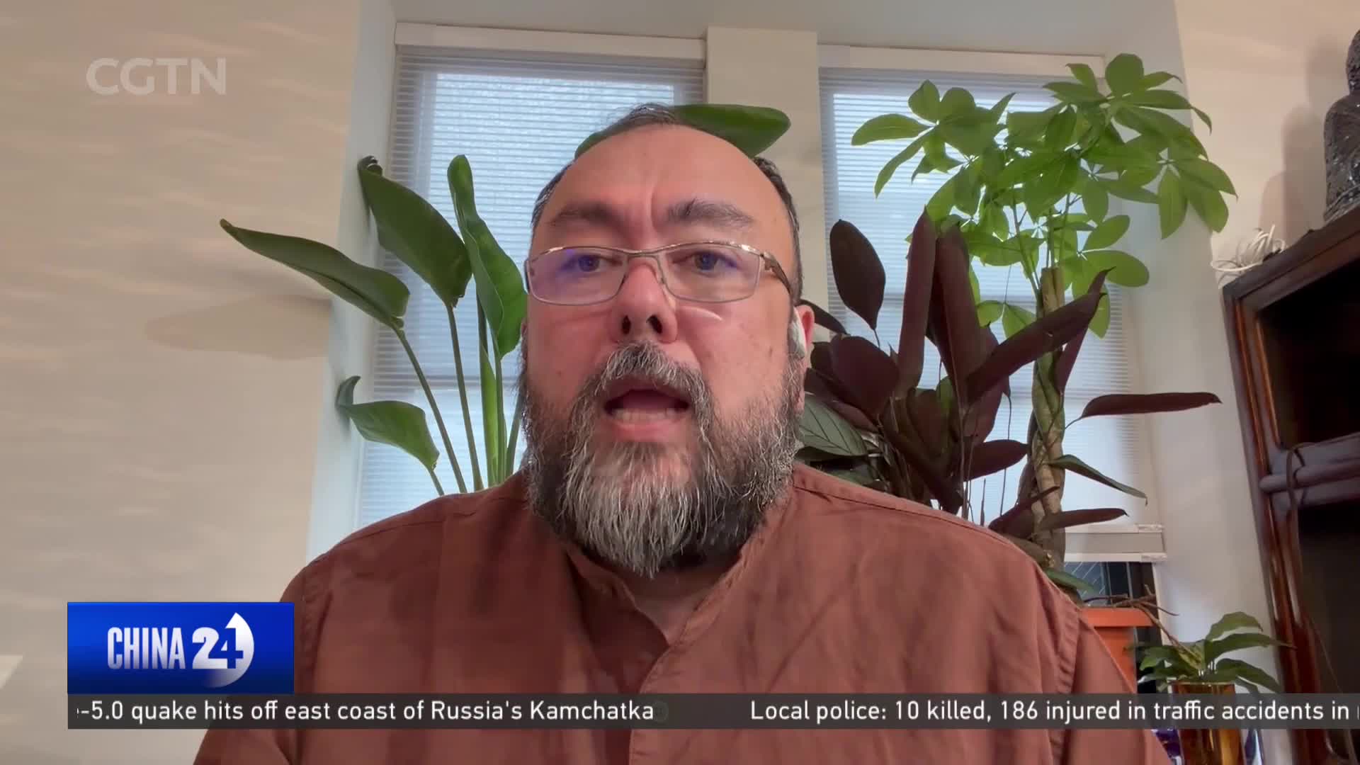 A man with glasses and a beard speaks directly to the camera, framed by lush green plants. Below him, the China 24 news ticker scrolls with updates on an earthquake and local police reports.