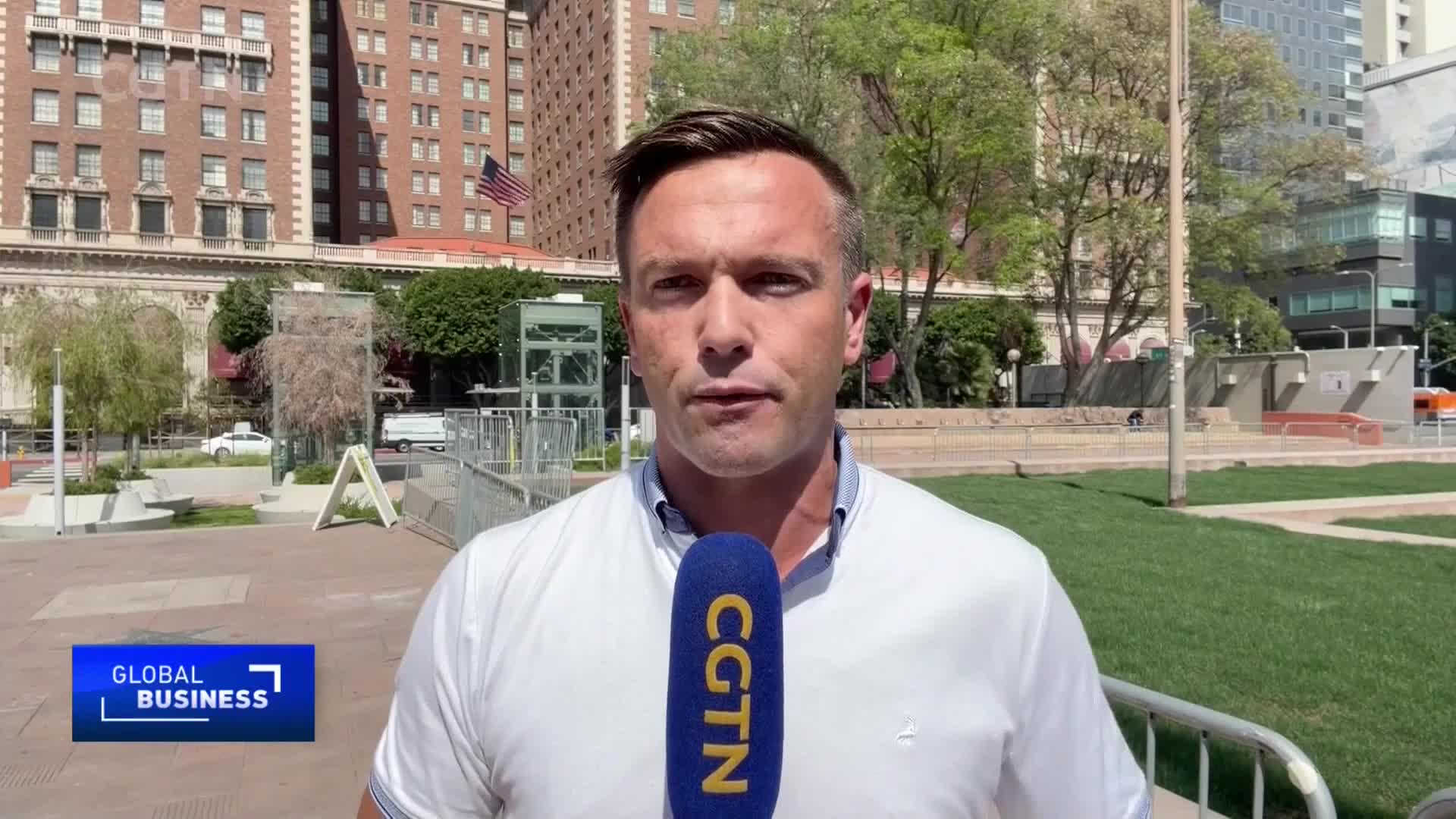 A man stands before a tall, brick building, holding a CGTN microphone. Behind him, a white car drives past a modern glass structure.