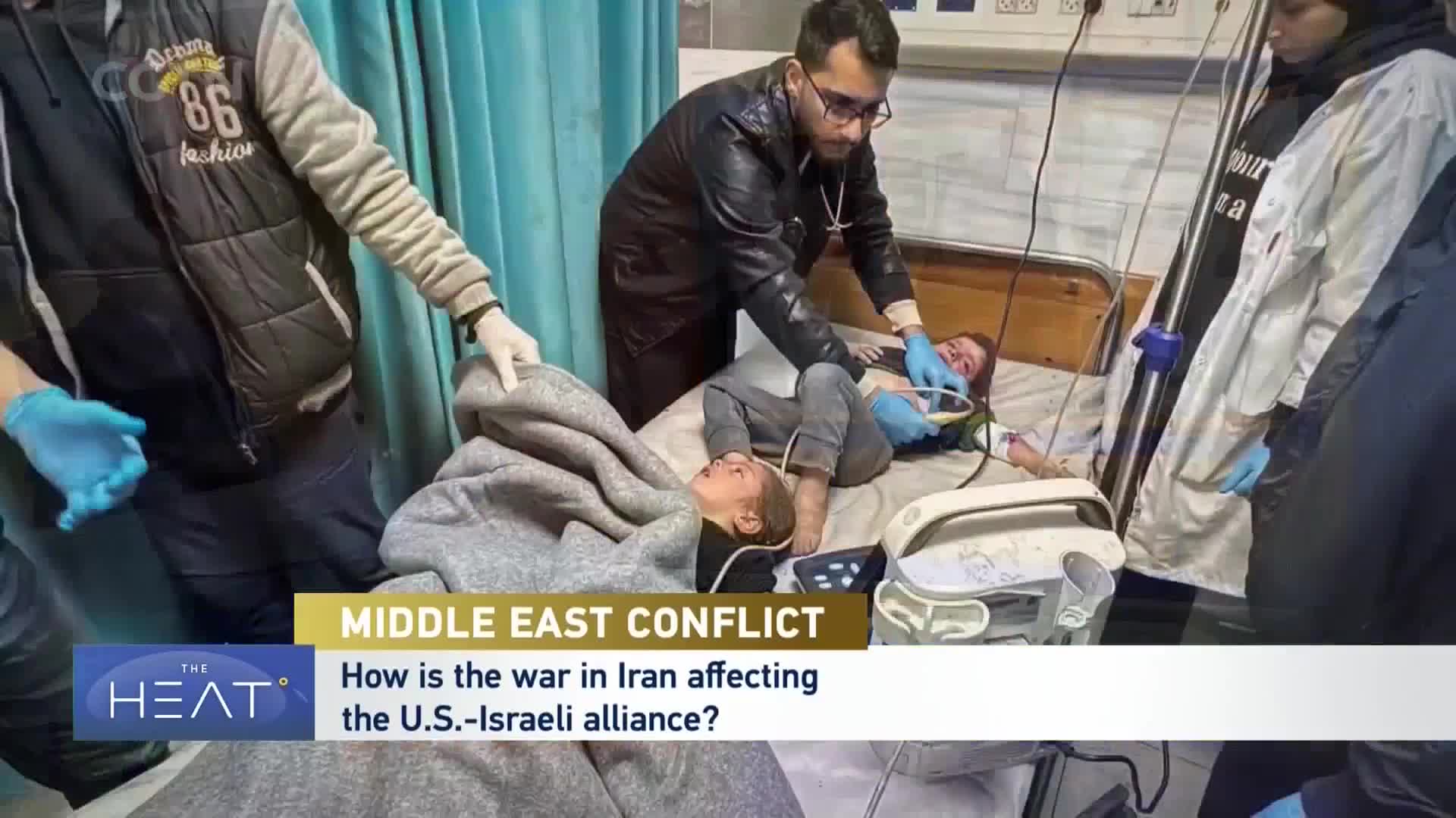 A medic, wearing glasses and a dark jacket, leans over a child lying on a hospital bed, attending to their face. Another child, wrapped in a gray blanket, rests on the same bed, their eyes closed.