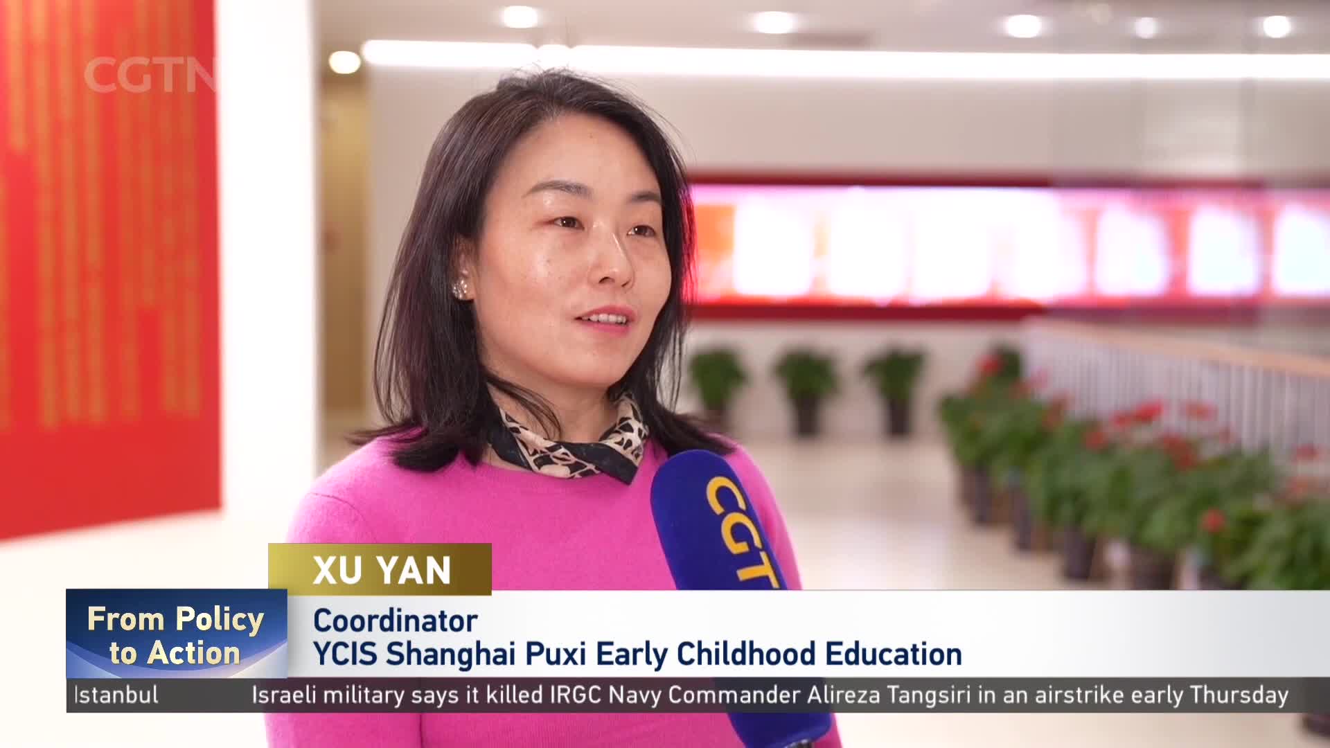 A woman in a bright pink sweater speaks into a microphone bearing the CGTN logo. Behind her, a hallway stretches into the distance, lined with potted plants and a red railing.