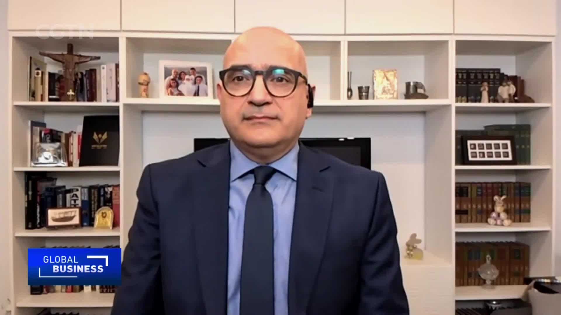 A man in a dark suit and tie looks directly ahead, his gaze steady. Behind him, shelves are filled with books and framed pictures, a subtle nod to his background and interests. The "GLOBAL BUSINESS" logo in the corner indicates this is a segment from CGTN.