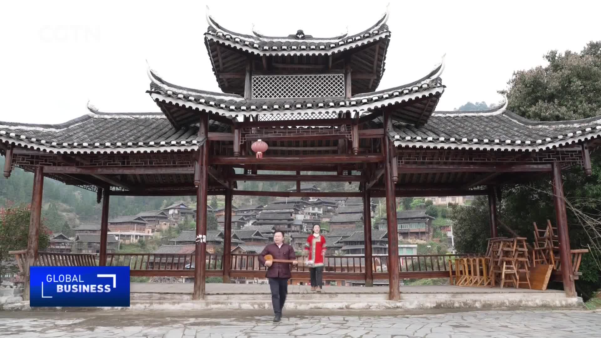 A man walks forward playing a stringed instrument while a woman stands behind him, both framed by a traditional wooden pavilion. Behind them, a village of wooden houses cascades down a hillside under a muted sky.