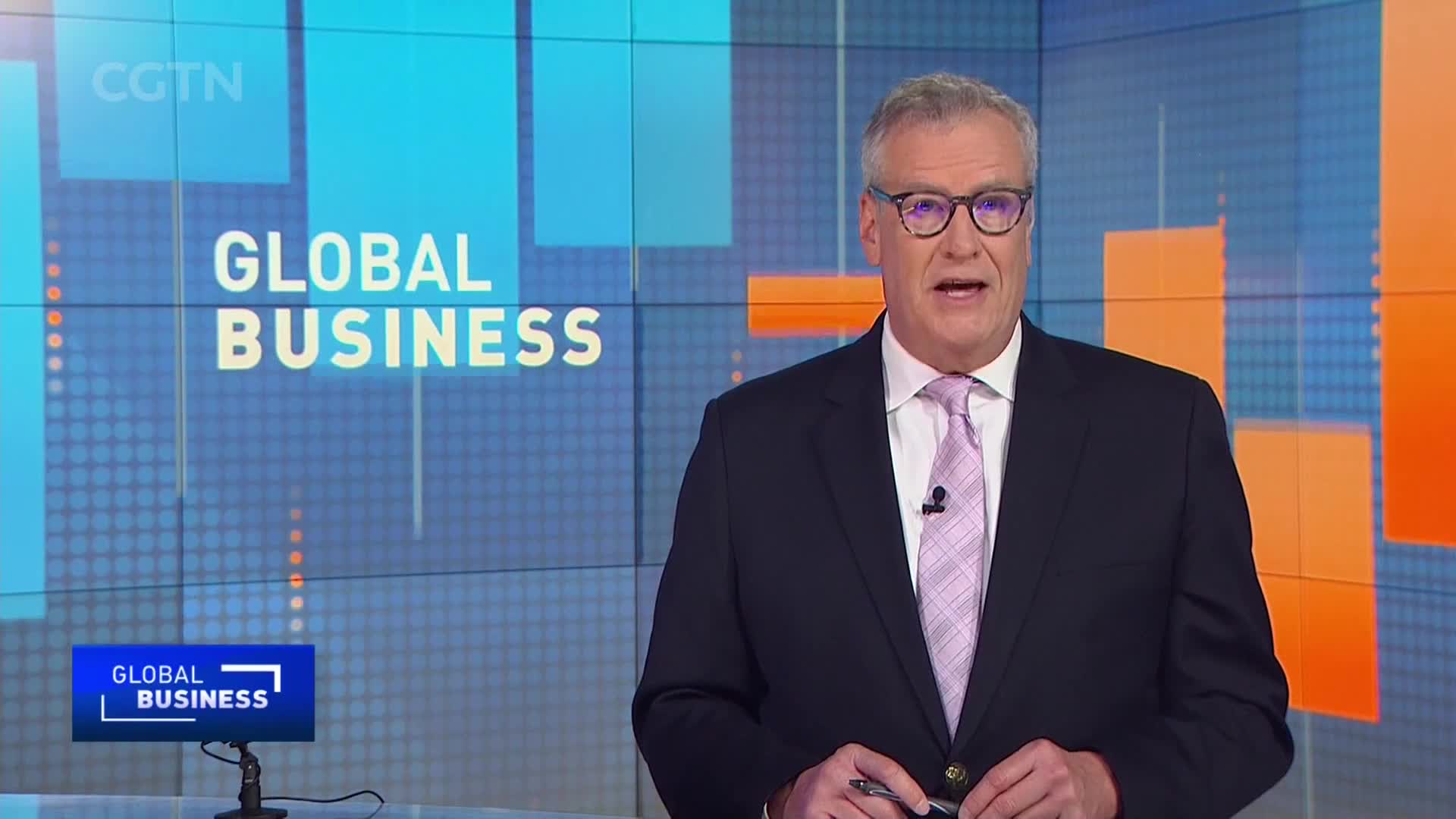 A man in a suit and tie stands before a screen displaying "GLOBAL BUSINESS" and the CGTN logo. He speaks directly to the camera, holding a pen in his right hand.