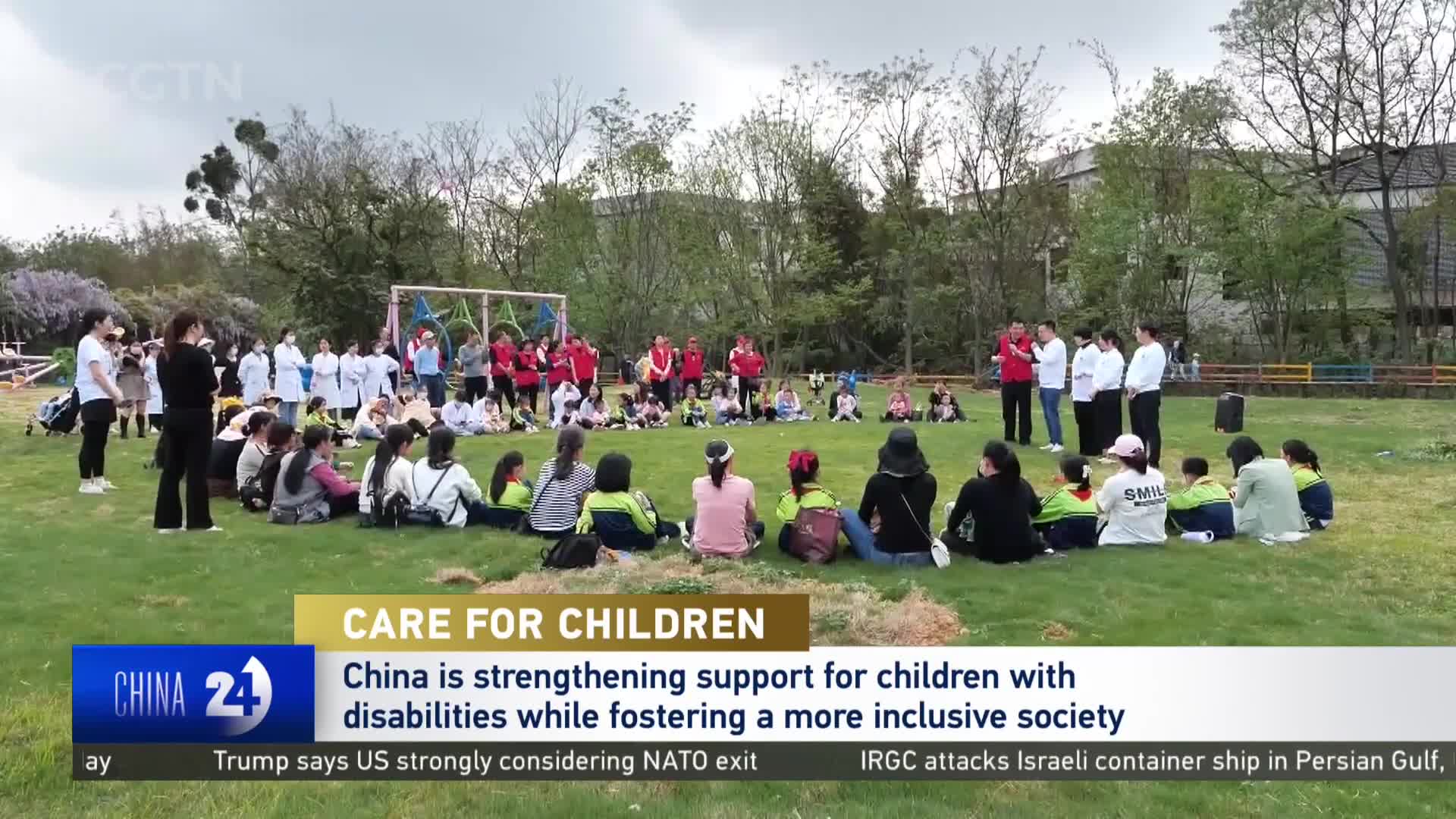 Adults in red shirts and white jackets gather in a circle on the grass, facing children seated on the ground. A group of women in white coats stands to the left, observing the scene. Adults in red shirts and white jackets gather in a circle on the grass, facing children seated on the ground. A group of women in white coats stands to the left, observing the scene.