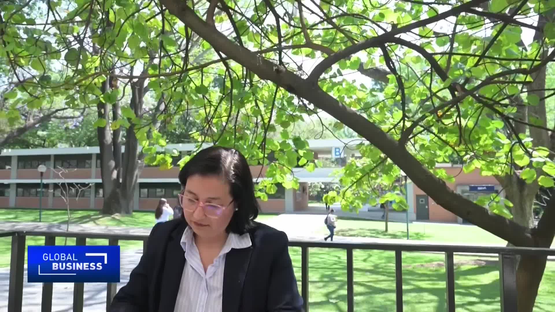 A woman in a dark suit speaks while looking down, framed by the dappled sunlight filtering through green leaves. In the background, a student walks across a grassy quad in front of a modern building. A woman in a dark suit speaks while looking down, framed by the dappled sunlight filtering through green leaves. In the background, a student walks across a grassy quad in front of a modern building.