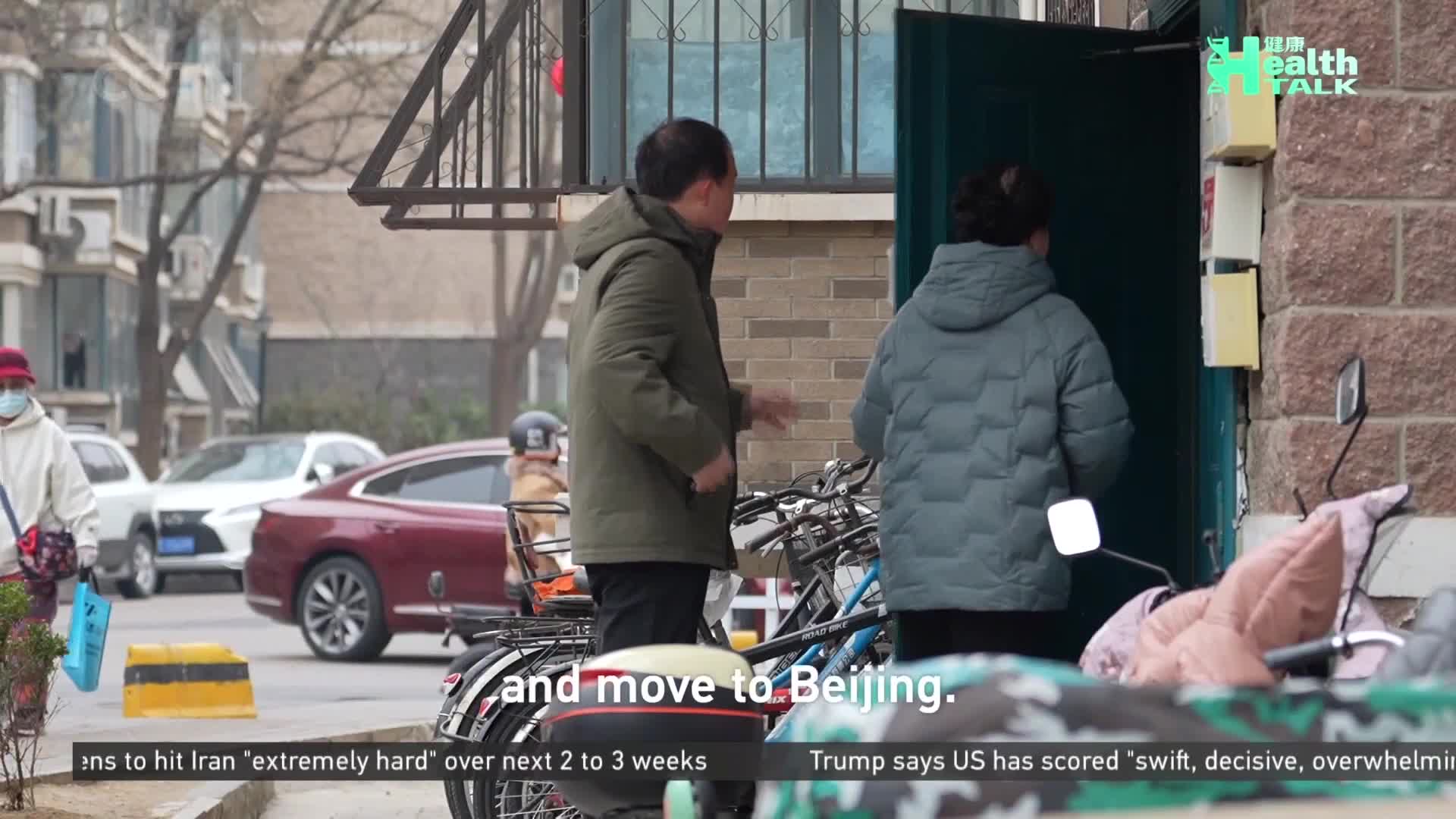 A man in a green jacket gestures as a woman in a puffy grey coat enters a doorway in Beijing. A woman with a red hat walks past parked cars in the background. A man in a green jacket gestures as a woman in a puffy grey coat enters a doorway in Beijing. A woman with a red hat walks past parked cars in the background.