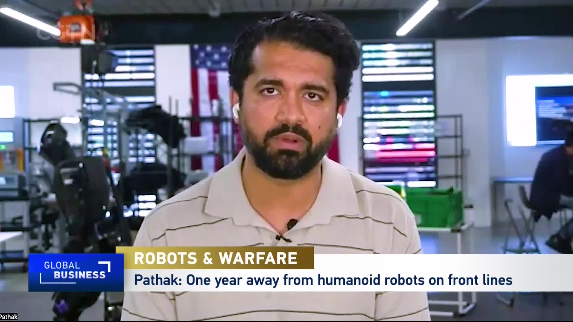 A man with a beard and earbuds speaks directly to the camera, a large American flag behind him. A robotic arm stands in the background, its metallic joints visible. A man with a beard and earbuds speaks directly to the camera, a large American flag behind him. A robotic arm stands in the background, its metallic joints visible.