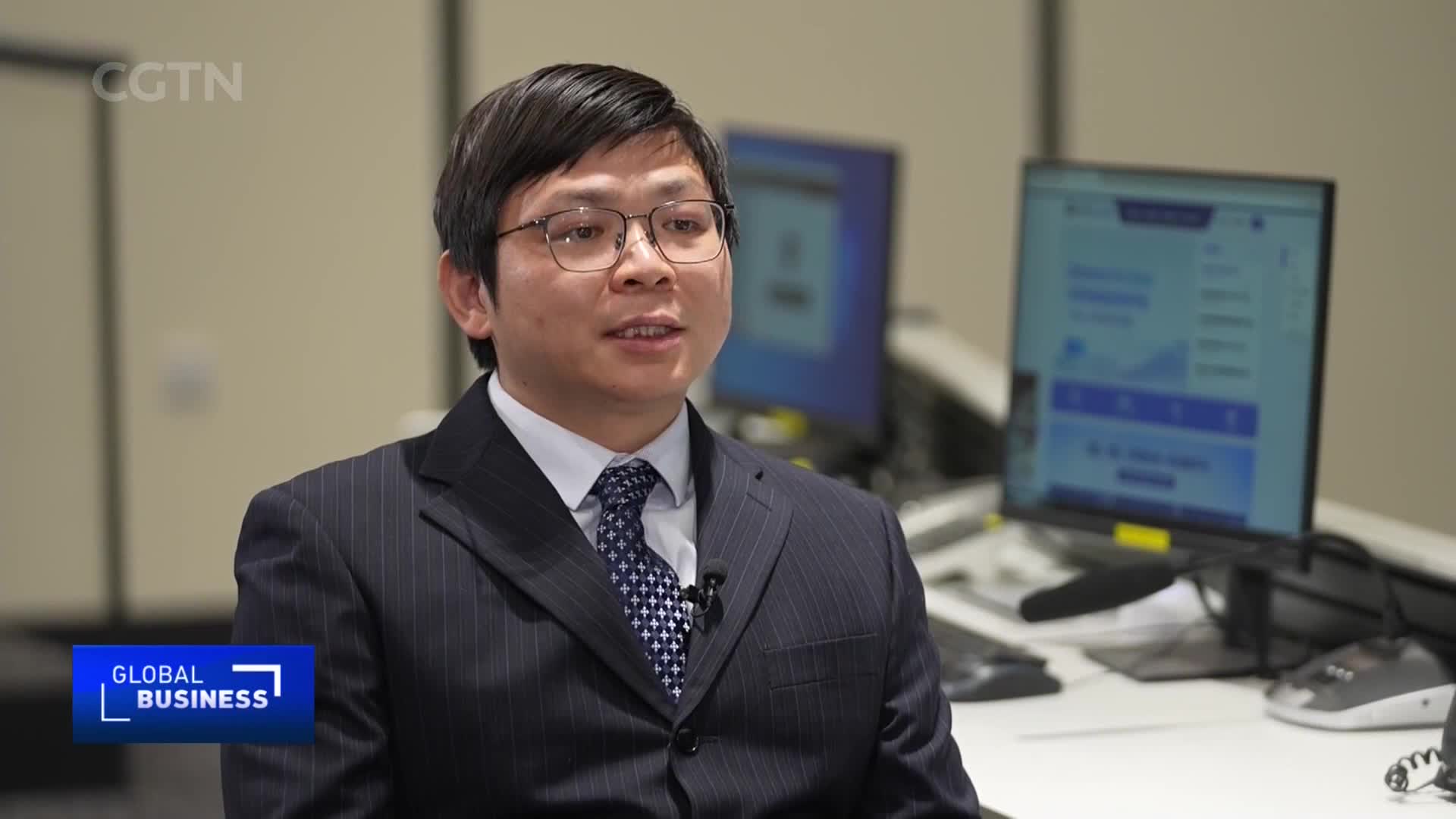 A man in a pinstripe suit and patterned tie speaks, his glasses reflecting the light. Behind him, computer monitors display what looks like data for CGTN's Global Business segment. A man in a pinstripe suit and patterned tie speaks, his glasses reflecting the light. Behind him, computer monitors display what looks like data for CGTN's Global Business segment.