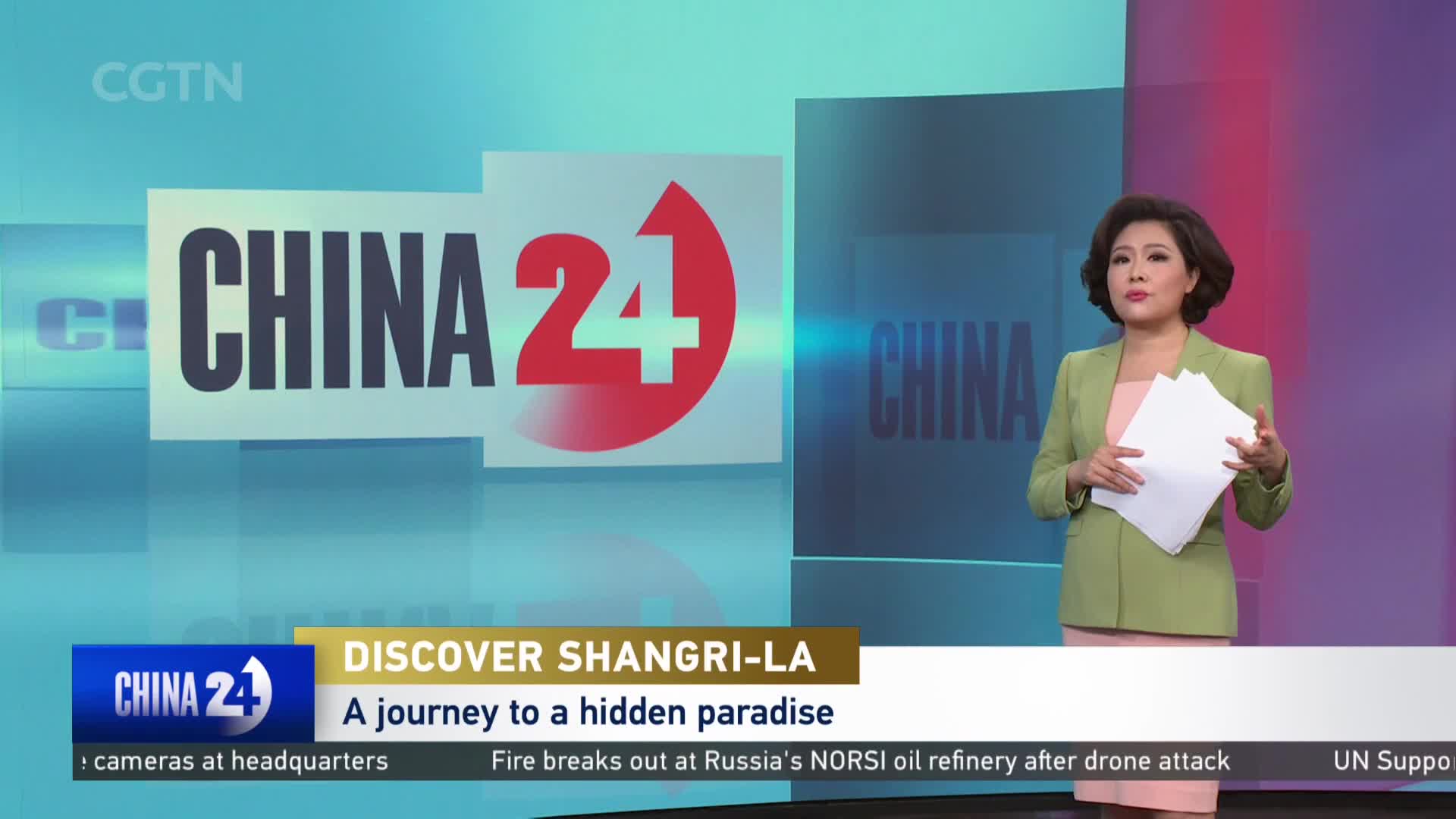 A news anchor stands before a large "CHINA 24" graphic, holding papers. Below, a ticker scrolls with headlines like "Discover Shangri-La" and "Fire breaks out at Russia's NORSI oil refinery."