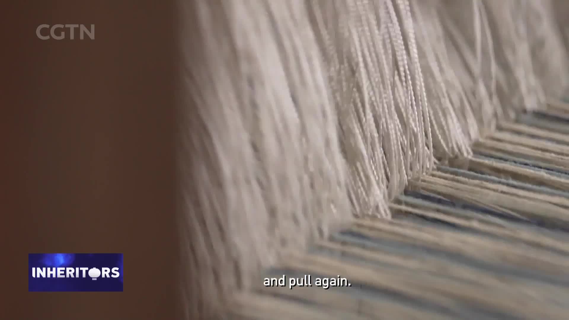 Threads of white and blue are being woven together on a loom. This is part of a CGTN report on traditional crafts in China, showcasing the "Inheritors" program.
