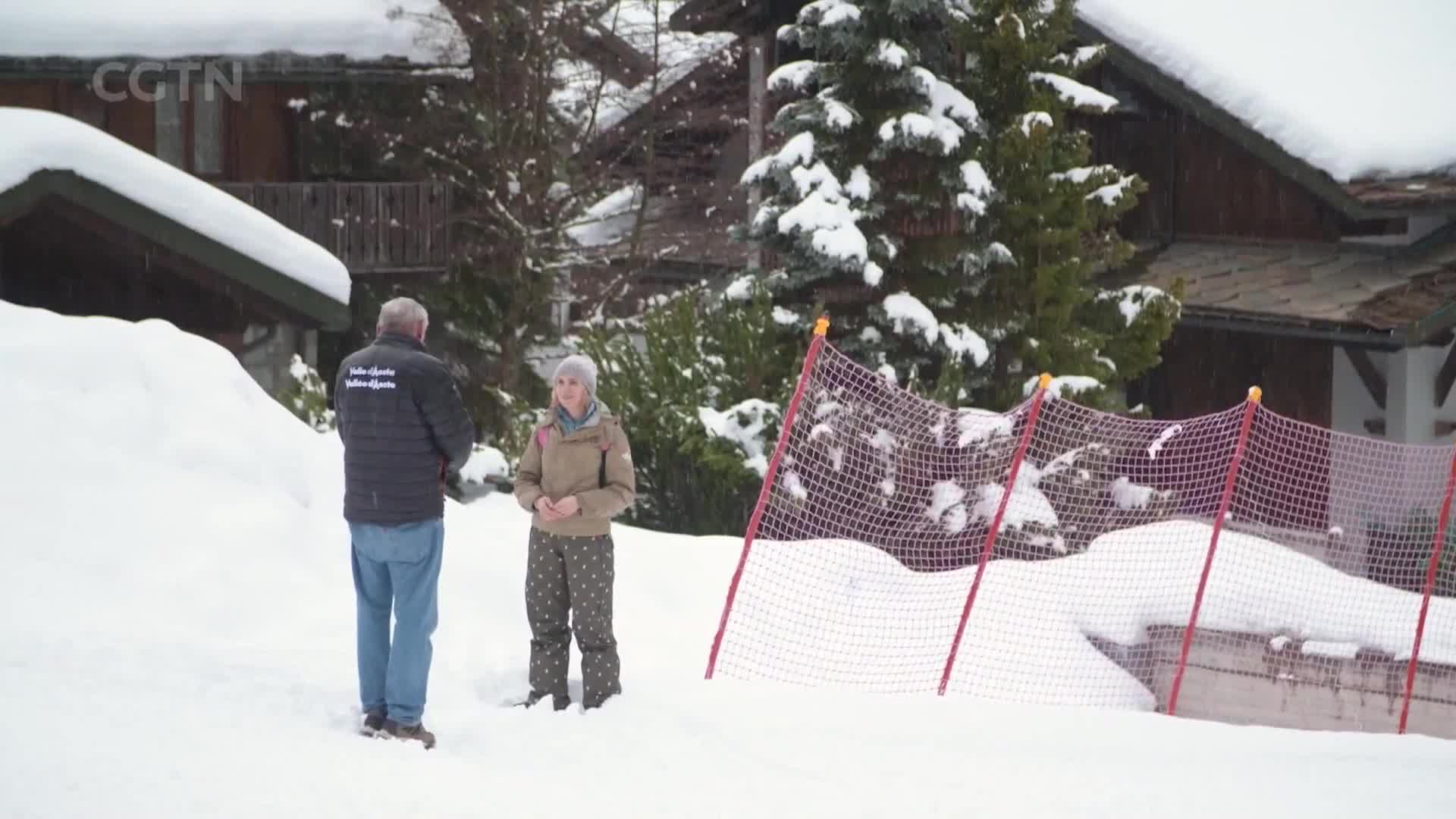 A man and a woman stand in the snow, talking near a red safety net. Snow-covered trees and wooden chalets surround them.