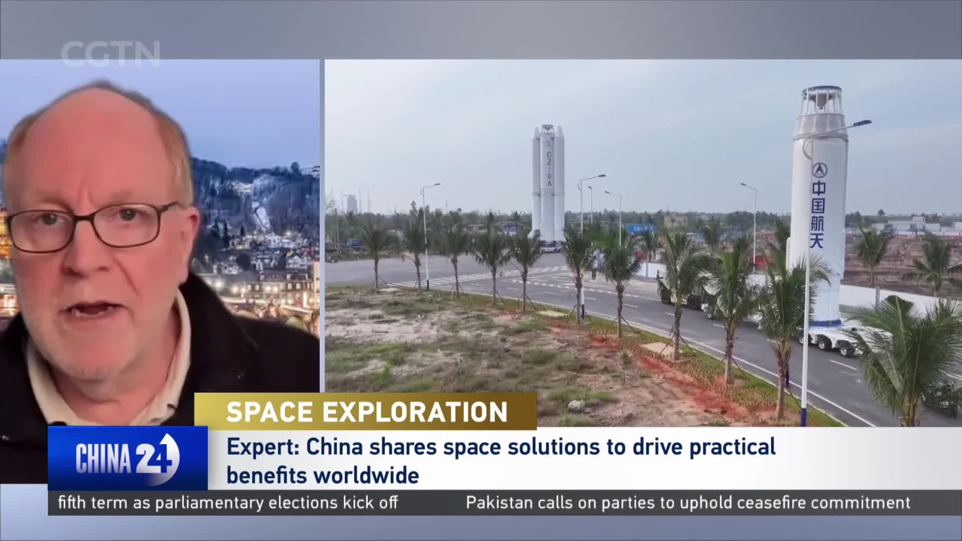A large rocket, emblazoned with Chinese characters, moves slowly down a road lined with palm trees. Trucks haul the towering structure past a launch facility, under a hazy sky.