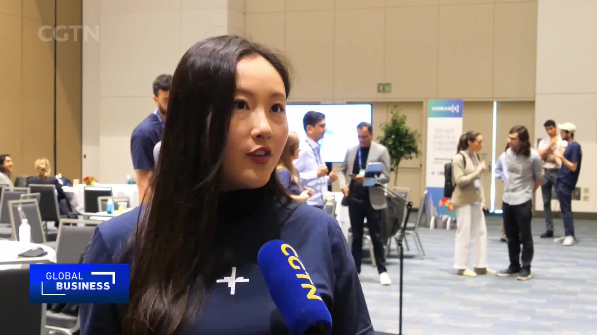 A young woman speaks into a CGTN microphone, her dark hair falling over her shoulder. In the background, people mingle near a presentation screen.