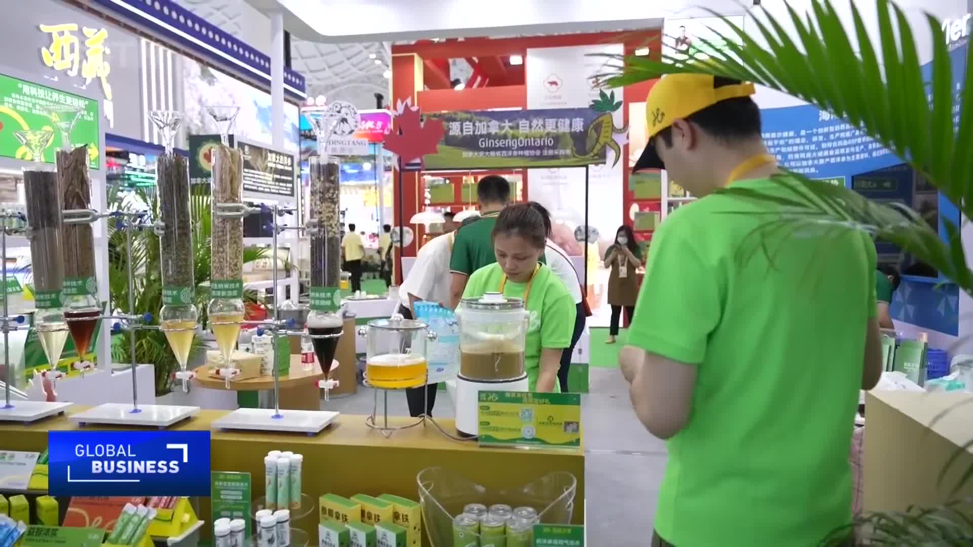 A woman in a bright green shirt operates a blending machine at a trade show booth in China. Behind her, a man in a yellow cap looks on as various ingredients are displayed in tall glass columns.