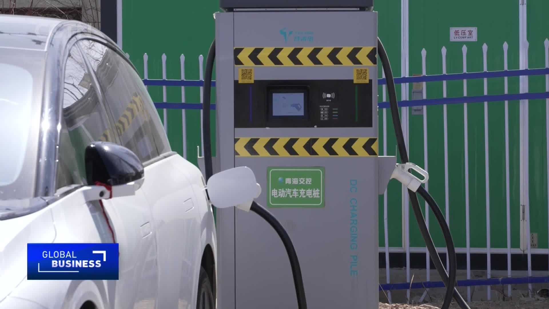 A white electric car is plugged into a charging station. The station displays Chinese characters and a "DC CHARGING PILE" label.