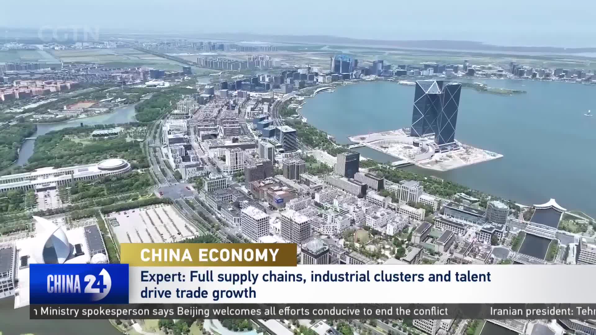 A vast cityscape unfolds, with modern buildings clustered around a wide, calm body of water. Two striking, diamond-shaped skyscrapers rise from an artificial island, their dark glass reflecting the sky.