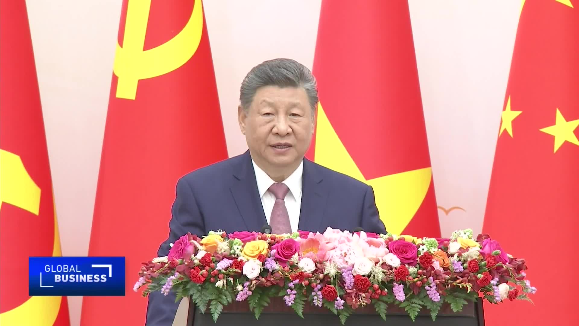Xi Jinping stands at a podium, speaking to an audience. Behind him, the red flags of China and the Communist Party are prominently displayed.
