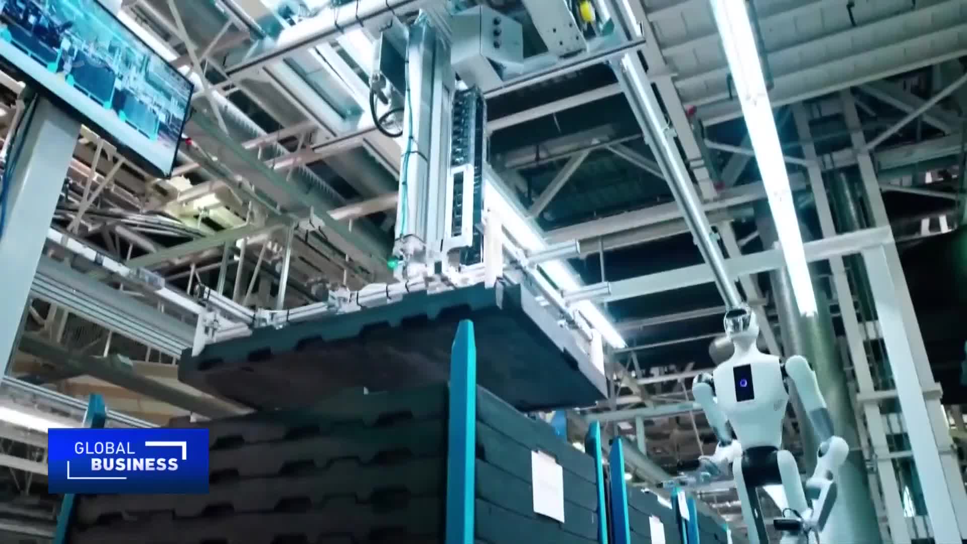 A robotic arm precisely lifts a large, dark panel, positioning it over stacks of similar items. Nearby, a humanoid robot stands ready, its chest displaying a glowing blue circle.