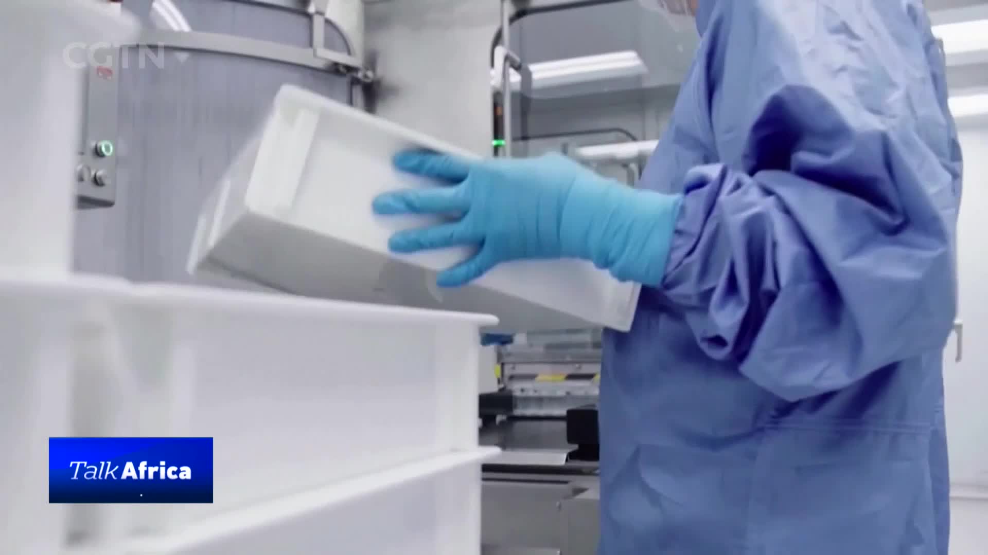 A gloved hand carefully places a white tray into another. The person wears a blue protective suit, typical for a cleanroom environment in China.
