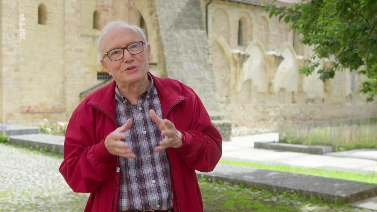 A man in a red jacket gestures with his hands as he speaks, the camera focused on him. Behind him, a stone building stands in the sunlight.
