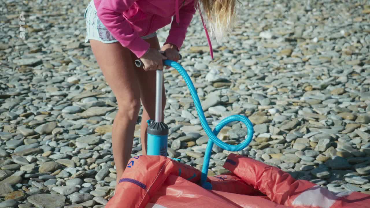 A woman in a pink hoodie pumps air into a red kite, the blue hose snaking across the stony beach. The logo of arte is visible in the corner.
