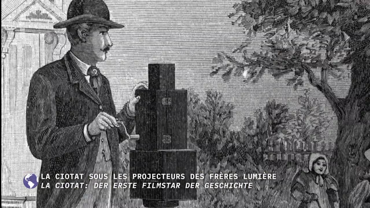A man in a suit and hat stands beside a tall, boxy camera, adjusting something with a small tool. In the background, a few people watch from beneath a large tree.
