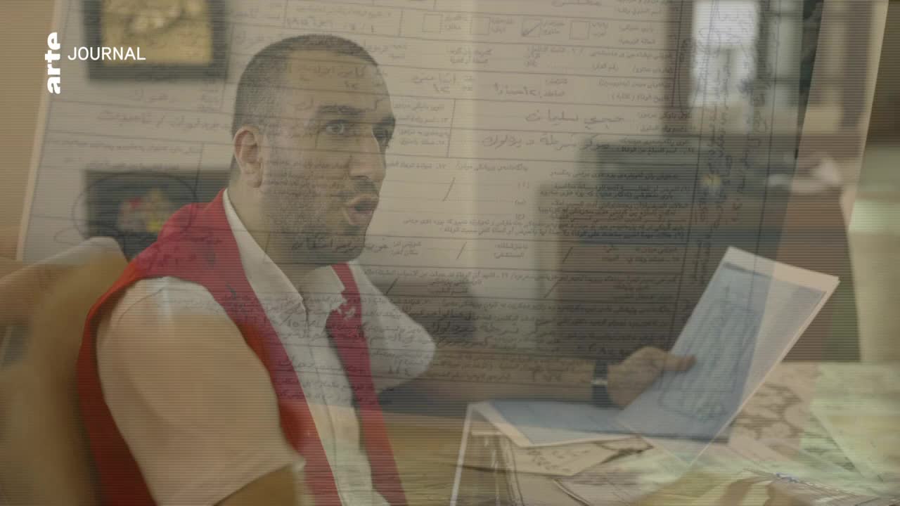 The man, wearing a red vest, gestures with one hand and holds a document in the other. Behind him, a superimposed document with handwritten text partially obscures the scene.
