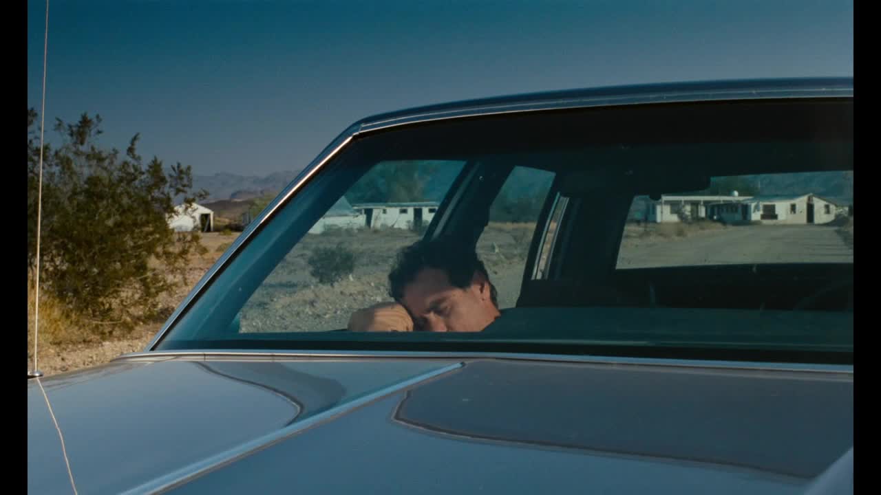 A man sleeps, his head resting against the car window. Beyond the vehicle, low buildings and dry scrub stretch towards the distant mountains.
