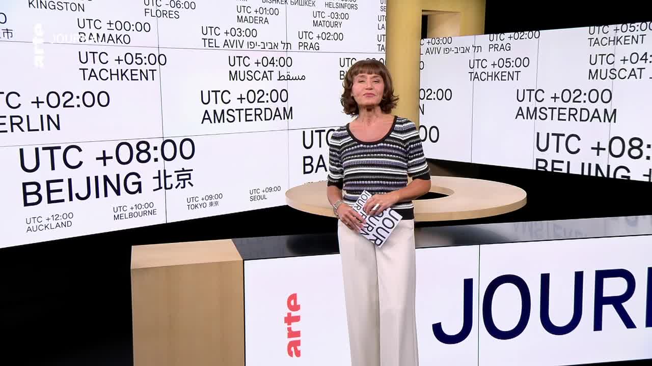 A woman in a striped top stands in front of a screen displaying world time zones, the arte logo visible in the corner. She holds a small notepad, ready to speak.
