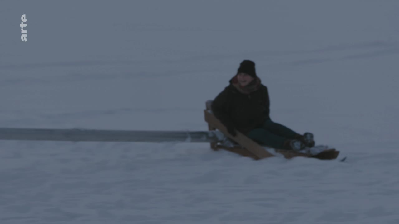 A person sits on a wooden sled, gliding across a snowy expanse. The sky is overcast, and the scene has a quiet, wintry feel.
