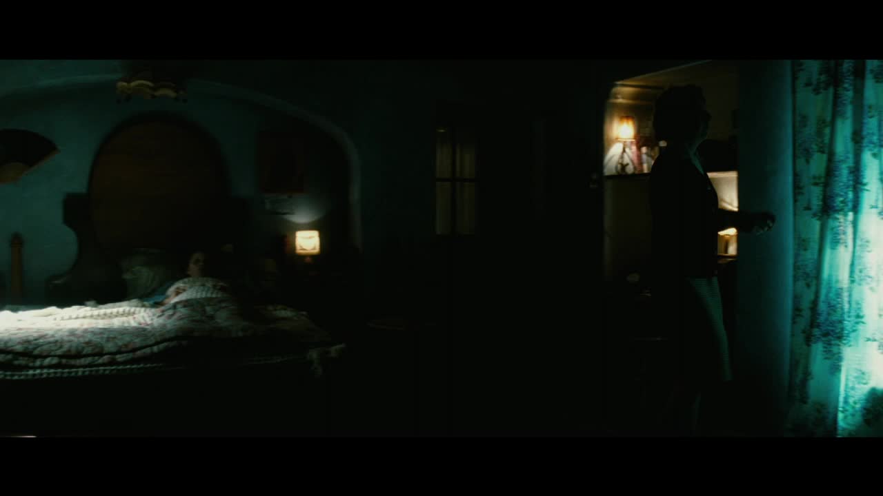 A figure stands silhouetted in the doorway, bathed in the soft glow of a bedside lamp. Across the room, someone lies still under a patterned quilt, the darkness punctuated by another small light.
