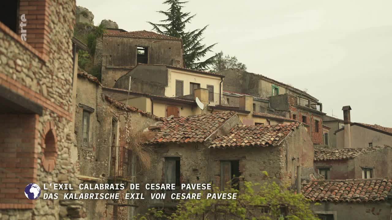 The camera pans across a cluster of weathered buildings, their terracotta roofs and crumbling facades suggesting age. A small tree stands tall against the overcast sky, its branches reaching towards the rooftops.
