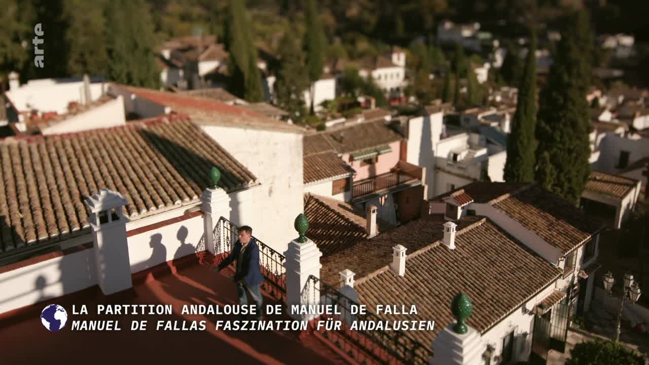 A man in a blue jacket walks across a red rooftop, casting a long shadow. The camera angle offers a view of the terracotta tile roofs and white buildings of the town below.
