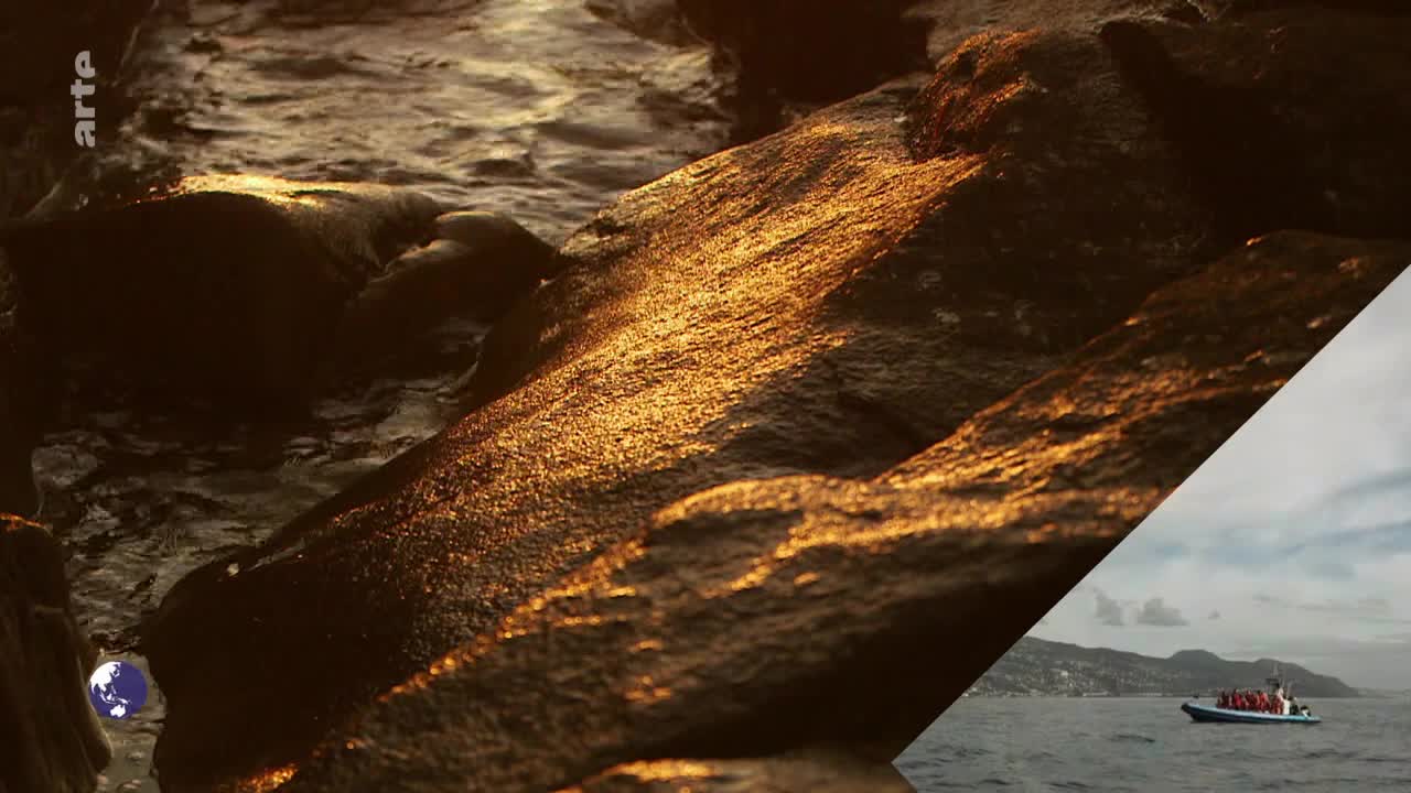 Golden light glints off the wet rocks as water gently laps against them. A boat filled with people moves across the water in the distance.
