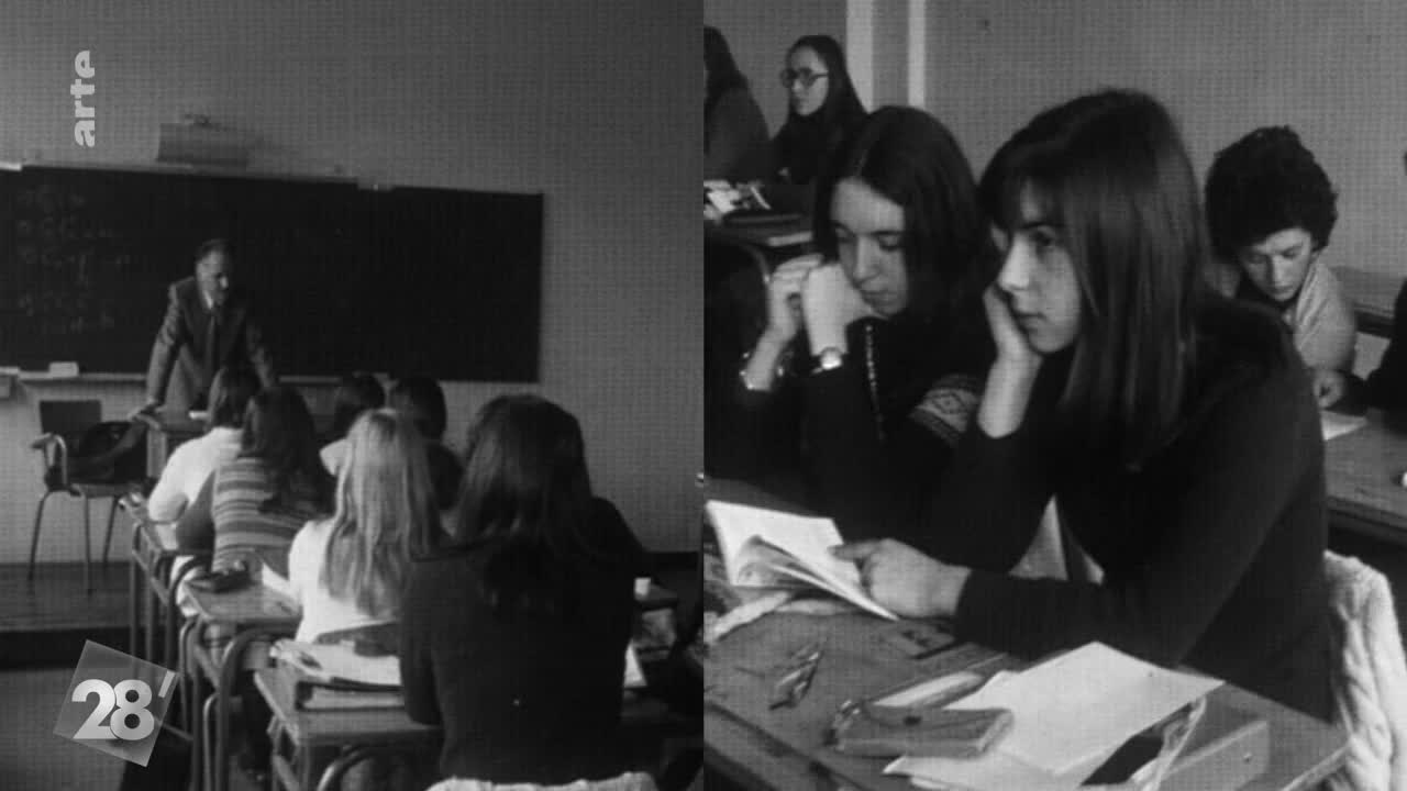 A professor in a suit lectures in front of a chalkboard, while students in the foreground listen intently. Across the room, several young women read and study, their faces illuminated by the classroom lights. 
