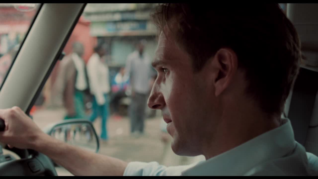 A man grips the steering wheel, his gaze fixed on something out of frame. The street outside is bustling, with a few figures blurred in the background.
