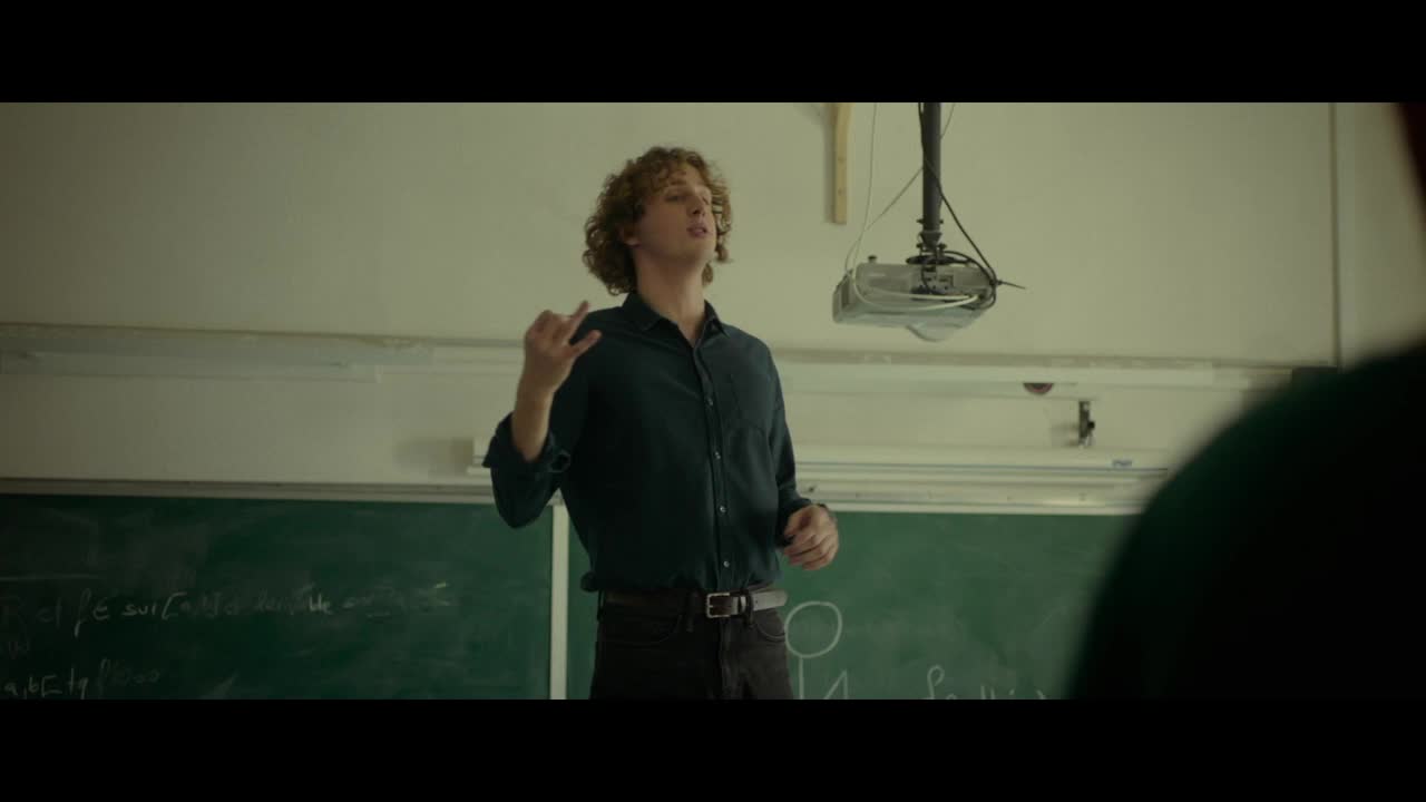 A young man with curly hair gestures with his hands, speaking in front of a chalkboard. Above him, a projector hangs, casting light onto the wall.
