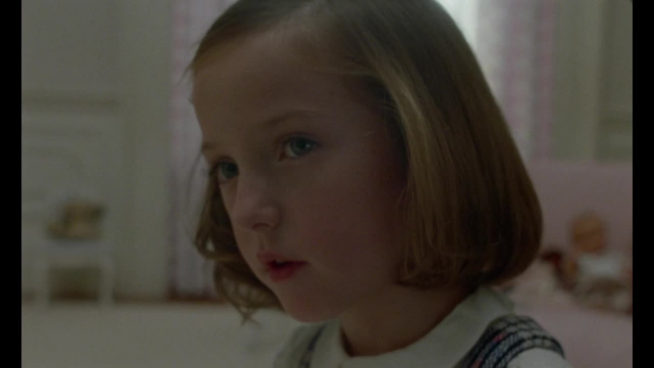 A young girl with pale skin and light hair looks directly ahead, her gaze steady. Her lips are slightly parted, as if she's about to speak.
