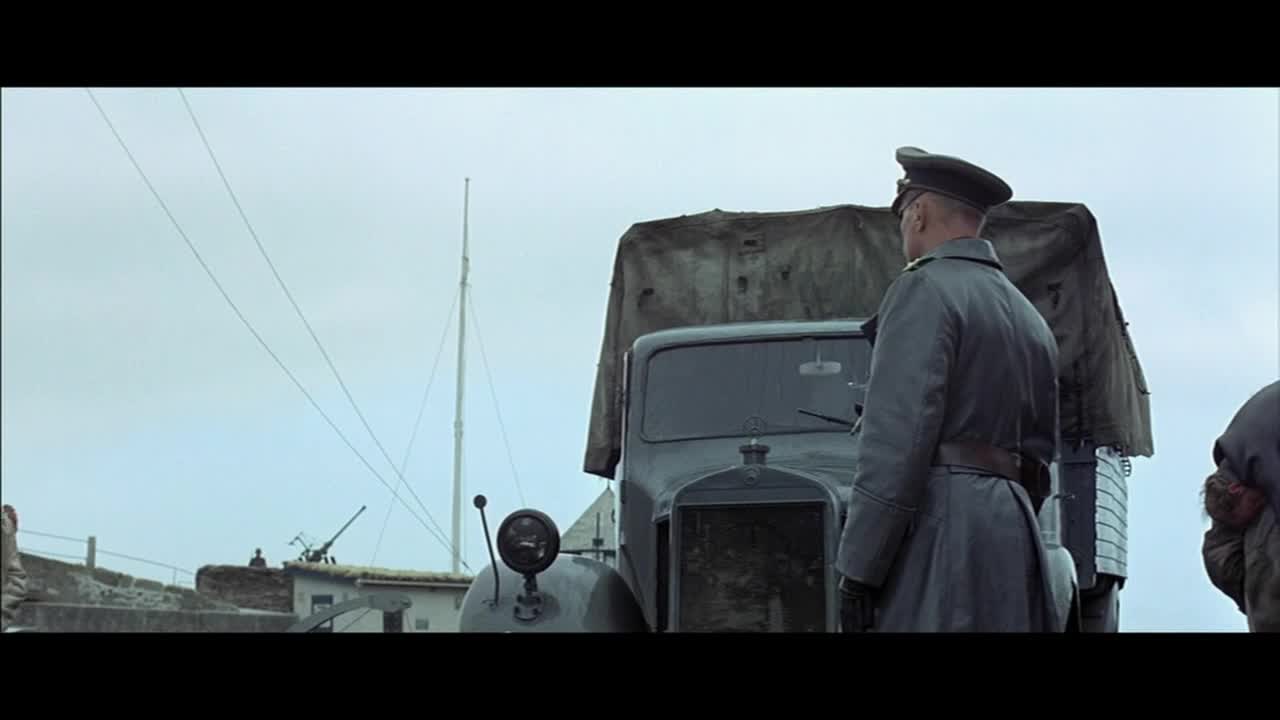 A man in a dark uniform stands facing away, next to a covered truck. In the distance, a small building with a mounted gun sits under a pale, overcast sky.

