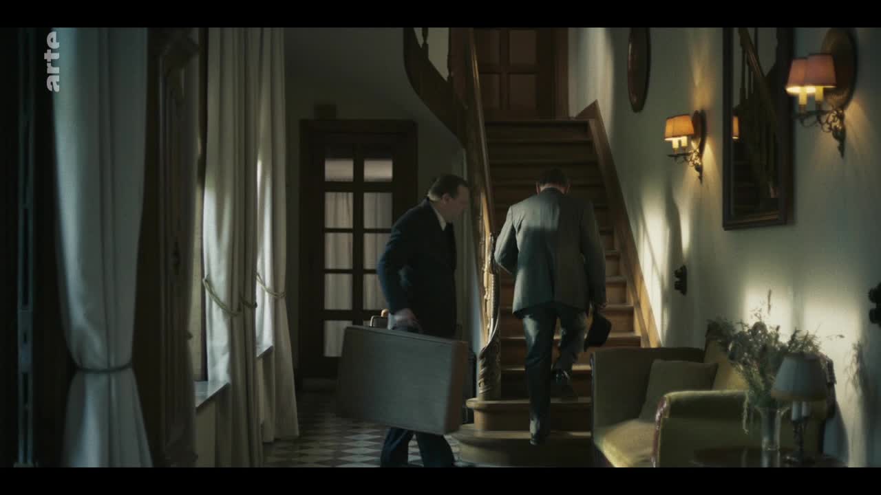 A man in a dark suit carries a suitcase, following another man up a wooden staircase. The light from a window illuminates the hallway, reflecting off the polished floor.
